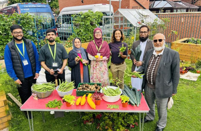 A group of people stands smiling behind a table filled with freshly harvested vegetables in a vibrant community garden.