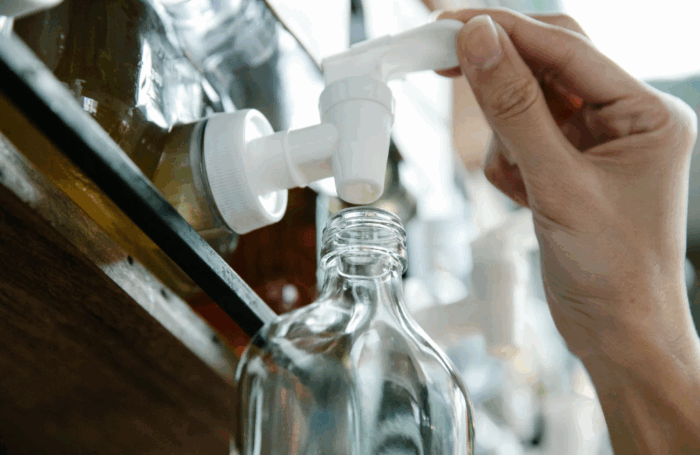 A person is dispensing liquid from a pump dispenser into a clear glass bottle.