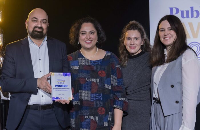 Four adults stand shoulder to shoulder at an awards event, smiling toward the camera. The person on the left holds a glass plaque reading “Public Finance Awards 2025 – Winner – Outstanding Procurement Initiative.” They are dressed in formal or business attire against a dark backdrop with event signage and stage lighting visible.