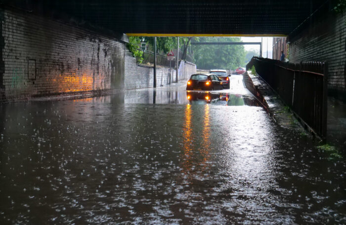 A flooded underpass with two cars driving through standing water on a rainy day. The brick walls and railway bridge above are wet, and reflections from the headlights are visible on the flooded road.
