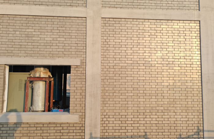 A beige brick building facade with concrete framing, featuring a small open window on the left that reveals industrial equipment and a wrapped cylindrical tank inside. Sunlight casts warm tones and faint shadows across the wall.