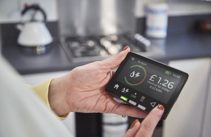 A person holding a home energy monitor in a kitchen. The screen shows the day’s electricity usage and cost, with £1.26 displayed.