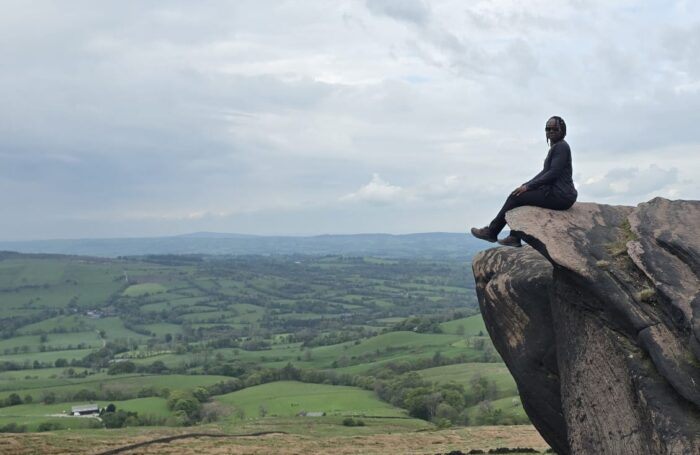 A person sits on the edge of a large rock outcrop overlooking rolling green countryside under dramatic grey clouds, taking in the expansive landscape.
