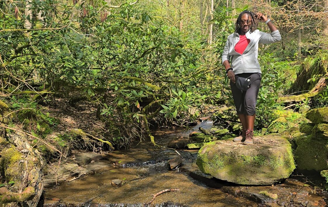 A person stands on a moss-covered rock in a shallow woodland stream surrounded by lush green trees and sunlight filtering through the forest.E